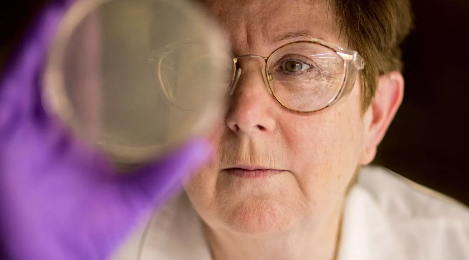 Close-up of a scientist wearing safety goggles and purple gloves inspecting a clear petri dish