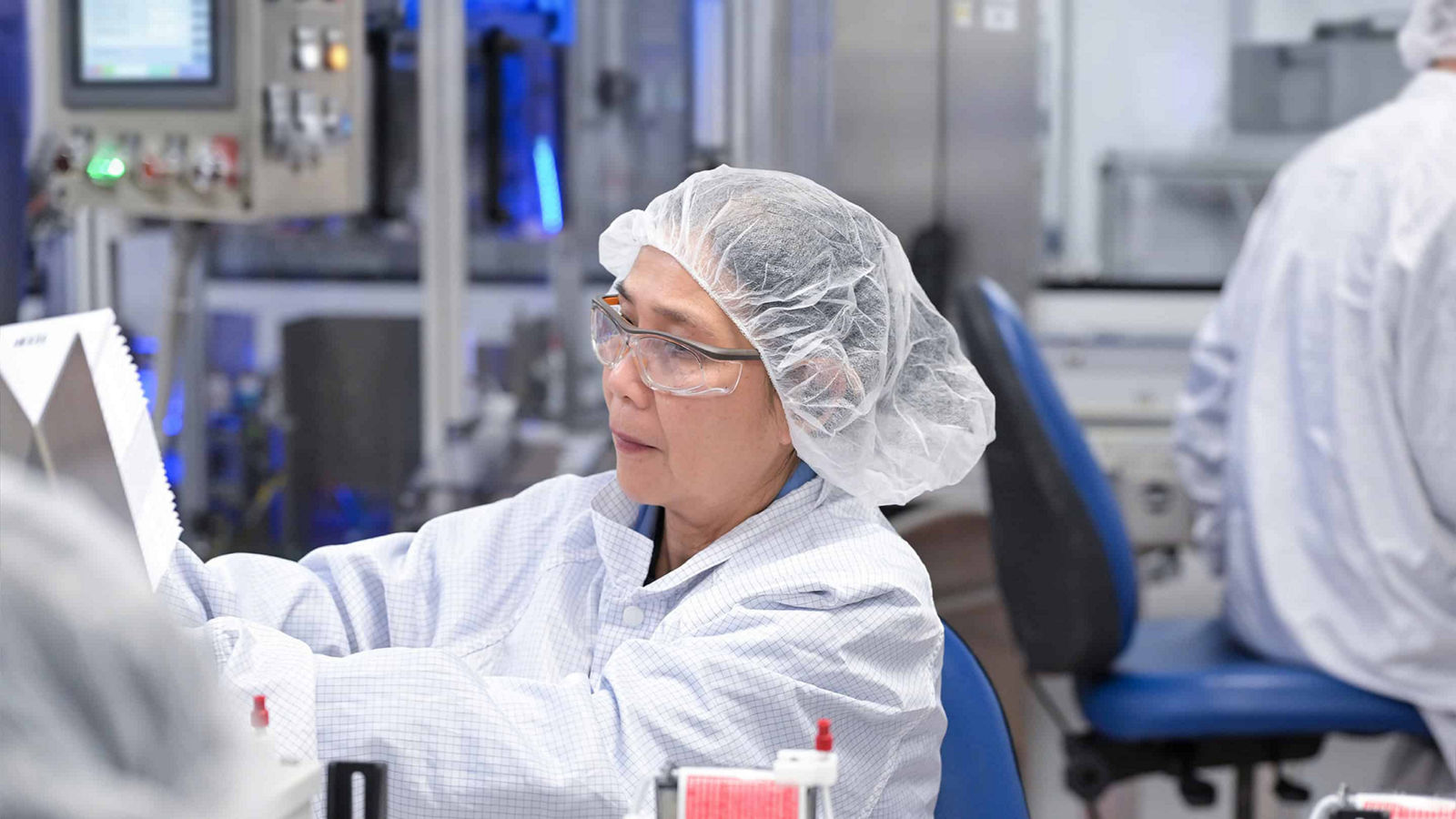 female employee in a white lab coat and a hair net wearing protective glasses working in a medication manufacturing facility