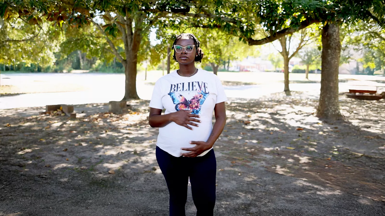 A pregnant woman in a white t-shirt and black pants stands in a park on a sunny day 