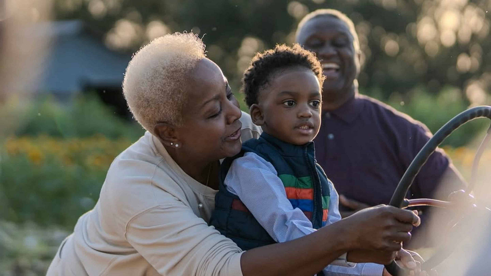 grandmother with rheumatoid arthritis helps her young grandson steer a tractor in a garden area at golden hour, as her smiling husband stands behind them