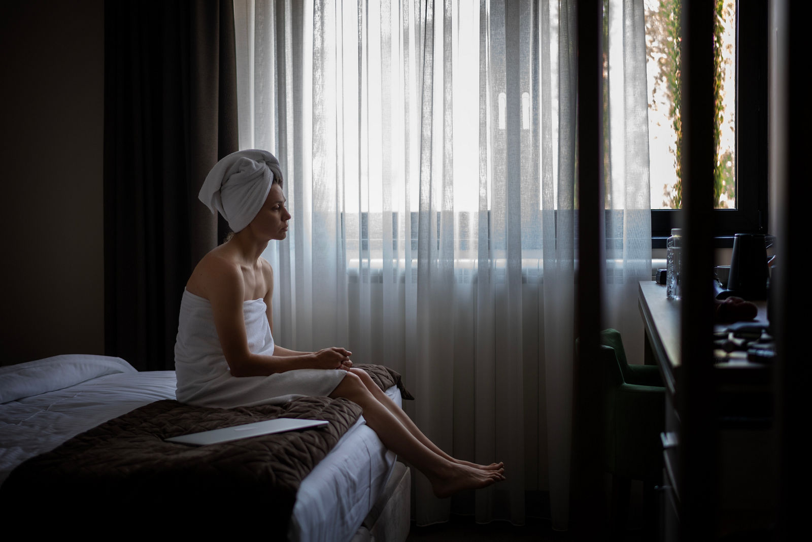 Sad young woman wrapped in towel after shower sitting on edge of bed in hotel room. 