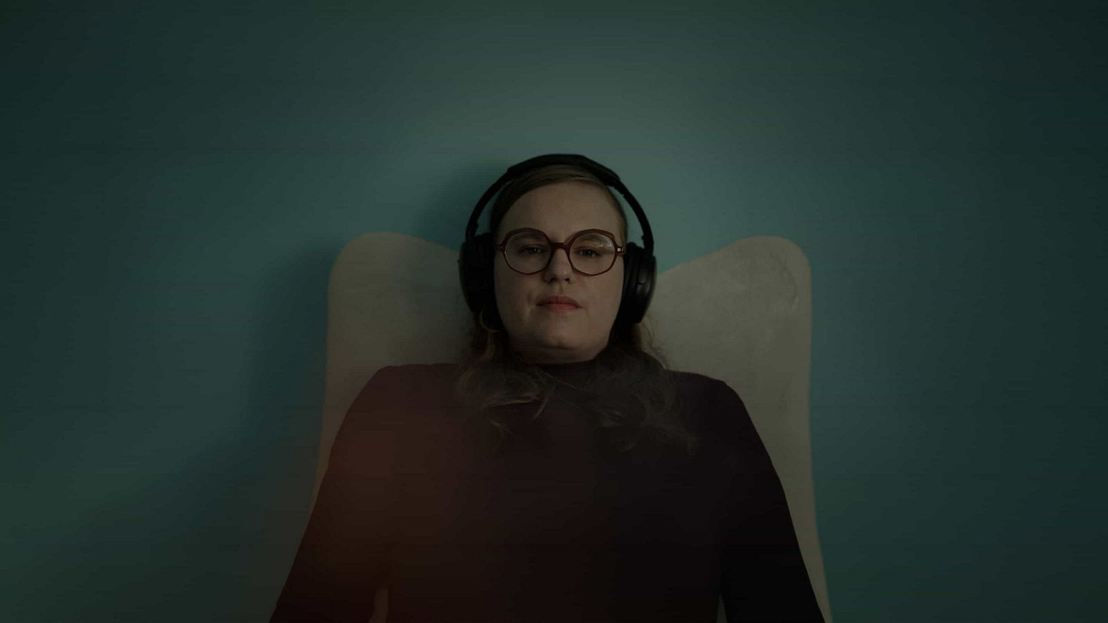 Morgane demonstrates how she copes with a migraine attack. She is reclining on pillows, wearing black over-ear headphones, in a calm, dimly lit environment